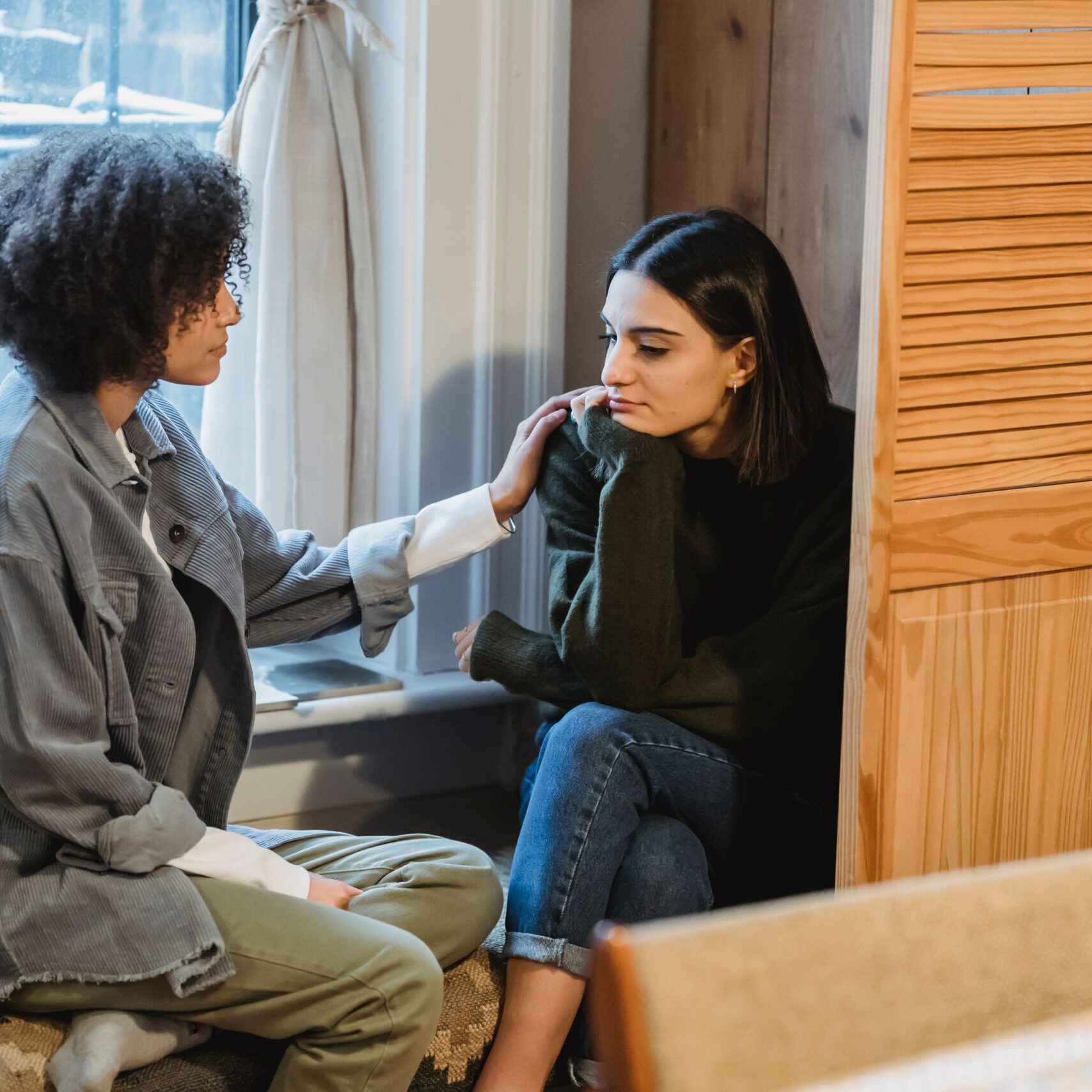 Calm young multiracial female best friends in casual clothes sitting near window and home and discussing discussing problems