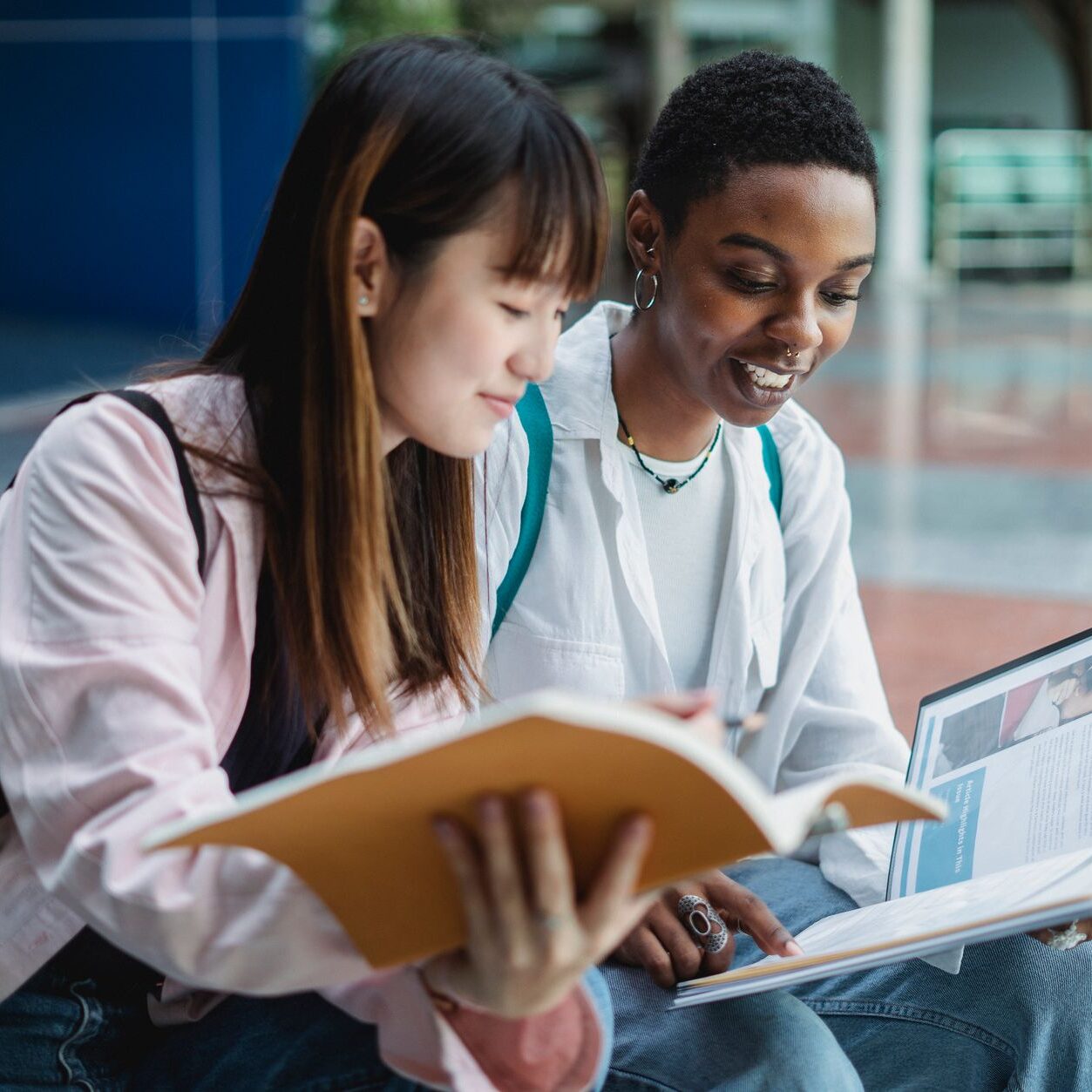 Young content multiethnic best female friends speaking while watching textbook on street stairs in city